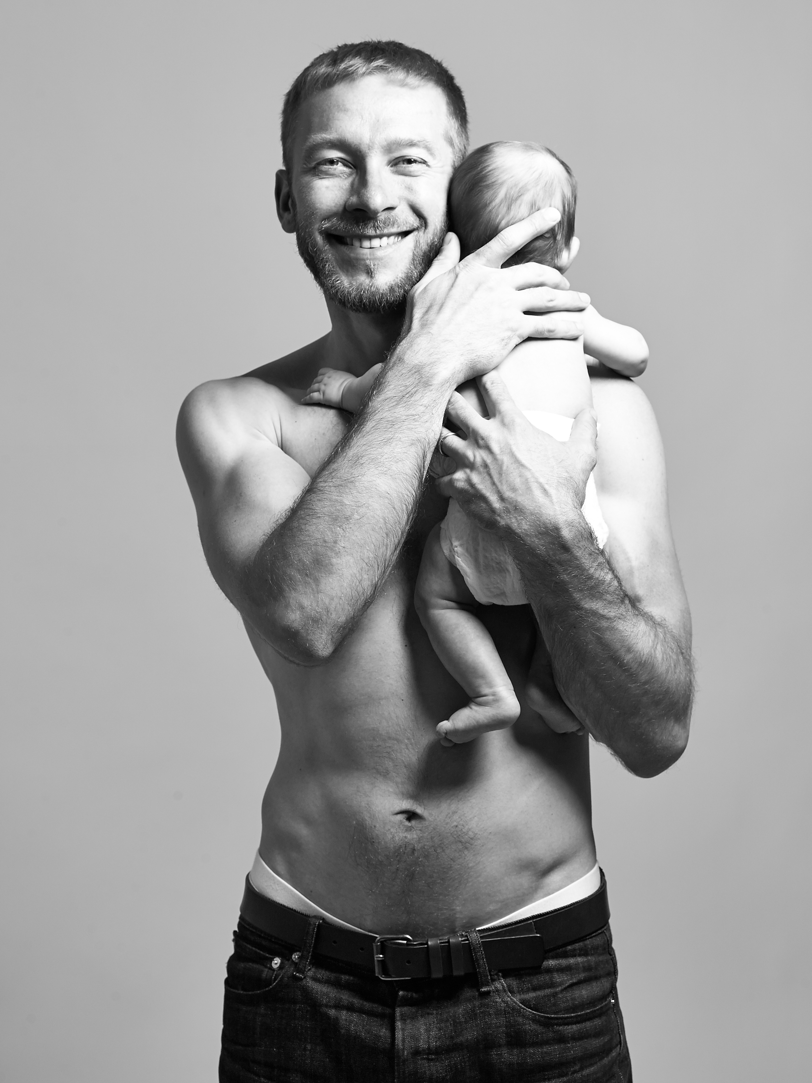 Cool guy with beard and smiling with naked upper body lovingly holding his baby daughter