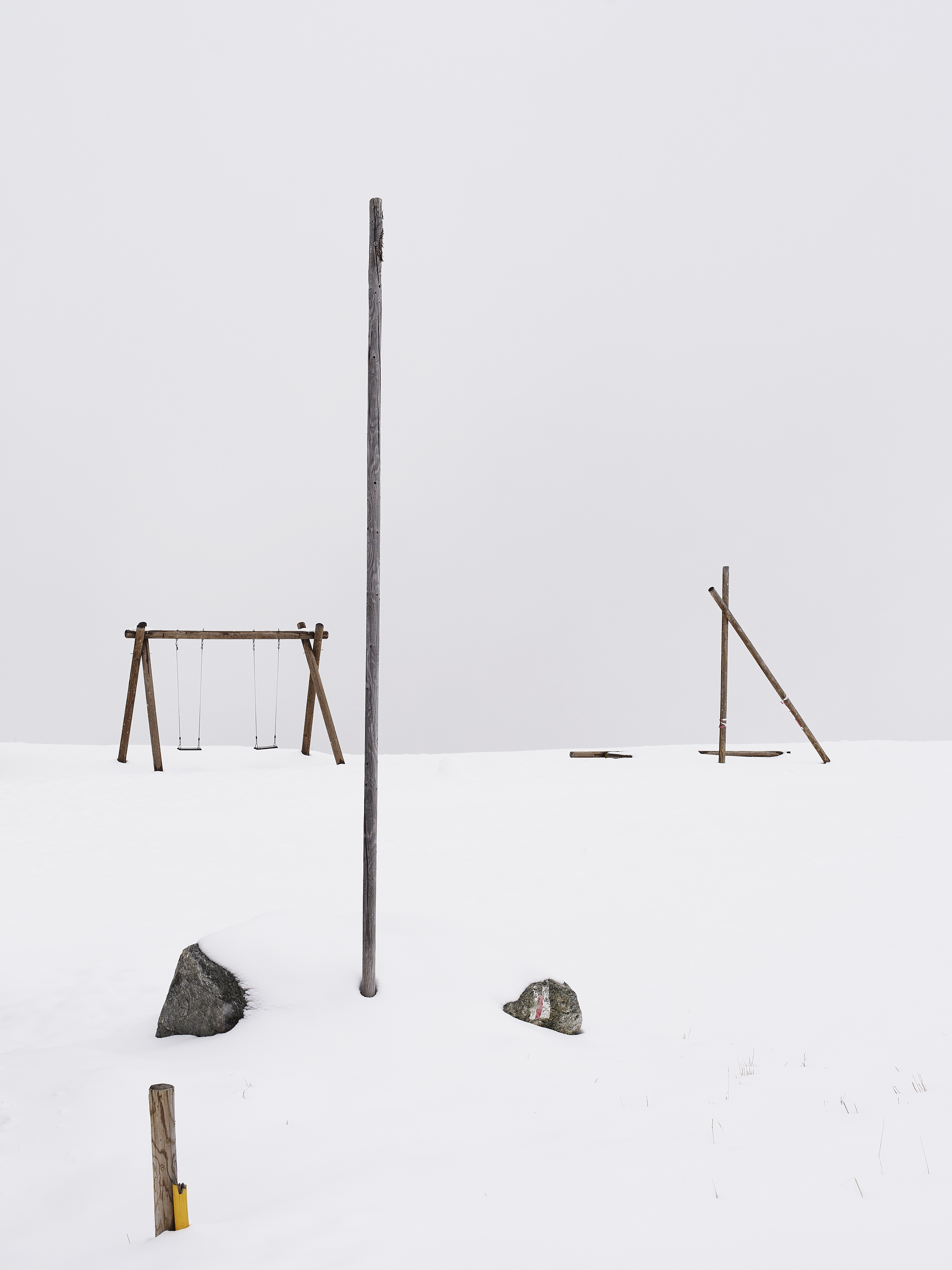 Minimalist photograph of some features of a wooden playground covered by snow in the foggy high Alps.